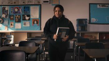 The Class TV show screenshot 2: A teenage girl in a dark sweatshirt walks into an empty classroom holding a notebook.