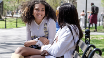 Two young women sit on a park bench. The woman at left has voluminous curly hair and is smiling at the women next to her, whose face we cannot see.