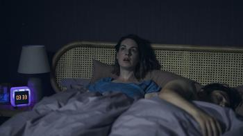 The Devil's Hour TV show screenshot 1: A woman sits awake in bed with a startled expression on her face. A man lays asleep next to her. The bedside cl