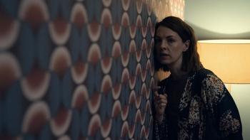The Devil's Hour TV show screenshot 3: A woman stands leaning closely against a patterned wallpapered wall, with one knuckle against it as if she is k