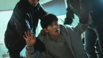 The Manipulated TV show screenshot: A young Korean man is held down by two other figures off-camera. His face is startled and arms up in surrender 