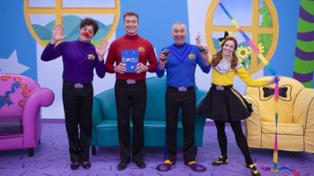 The Wiggles World TV show screenshot 1: Four Wiggles dressed in colored outfits stand in a living room with colored couches. 