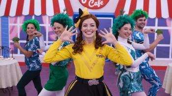 The Wiggles World TV show screenshot 2: White woman dressed in yellow front and center with hands open by her sides. Background actors in green.