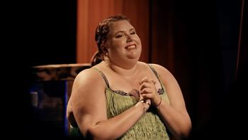 Thick Skin TV show screenshot 3: A smiling brunette woman in a green fringed dress clasps her hands at her chest and smiles.