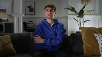 Thirst Trap TV show screenshot 1: William White sits on a dark grey couch. He wears a royal blue sweatshirt and smiles with his arms crossed