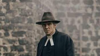 To Cook a Bear TV show screenshot 1: Actor Gustaf Skarsgård dressed in a dark hat, coat and white collar (period piece attire) for a Swedish priest