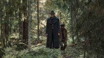 To Cook a Bear TV show screenshot 3: Two men, one tall, and one shorter, walk through a dense forest wearing dark clothing atop a forest floor of moss