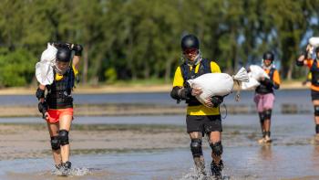 Trainer Games: White men and women run along a beach wearing helmets and elbow and knee pads carrying sand bags.