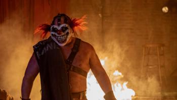Twisted Metal: Sweet Tooth is shown from the waist up, silhouetted against a roaring fire. His hair is wild and red and he wears a menacing clown mask.