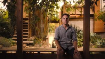 Twisted Yoga TV show screenshot: A man is shown sitting on a bench in front of a window with bamboo and other nature growing behind him.