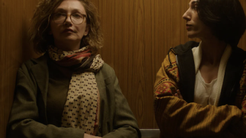 Unconditional TV show screenshot 3: Two people in an elevator, one in glasses and scarf, the other in a patterned jacket.
