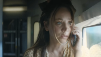 Vanished TV show screenshot 3: Cuoco holds a phone in her hands. She stands in a train cart. She wears a print dress and green ribbon in her hair.