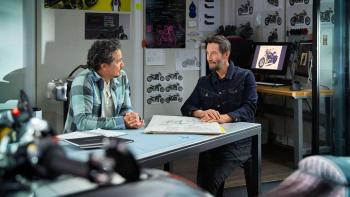Gard Hollinger and Keanu Reeves sit at a table in a workshop. They are surrounded by motorcycle posters and images.