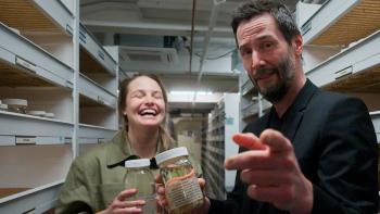 Keanu Reeves points at a camera while holding a jar. A woman in the background laughs, holding a jar as well.