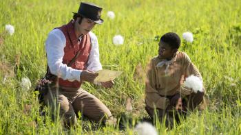 Tom Ellis and Eddie Karanja. Ellis wears a red waistcoat, tan pants, and brown hat. Karanja wears a tan shirt and brown pants.