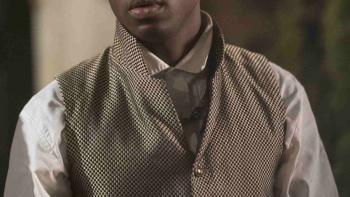 Ernest Kingsley Junior wears a checker patterned waistcoat over a white dress shirt. A gold chain hangs off one of the buttons.