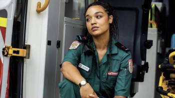 Watching You TV show screenshot 1: Aisha Dee sits on the back of an ambulance. She wears a dark green uniform with patches on the shoulders.