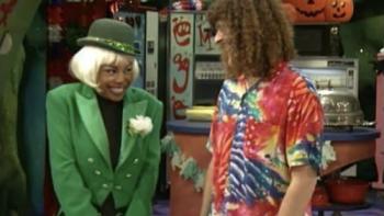 A giggling woman in a green coat and white wig talks with a Hawaiian-shirt clad Weird Al.