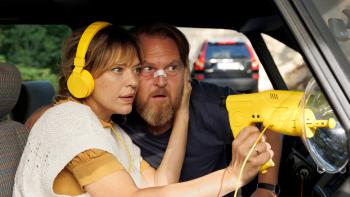 Heike Makatsch holds a bright yellow listening device as Axel Stein presses his heads up against hers. They are in a car.