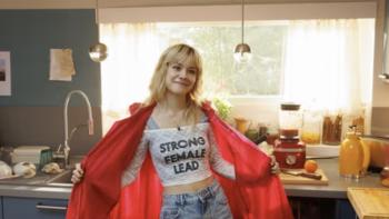 Lea Drinda stands in a kitchen. She holds open a red cape, revealing a shirt that reads “Strong Female Lead.”