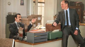 White House Plumbers TV show: Liddy sits behind a desk in an office with 1960s-era furnishings; Hunt sits on the desk, looking elated