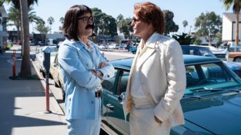 White House Plumbers TV show: Liddy and Hunt, in 1960s suits, stand in front of a vintage car on a street. Liddy wears a powder blue suit and black Prince Valiant-style wig; Hunt wears a beige suit and a rumpled orange wig