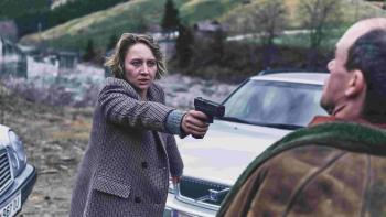 A woman with short blond hair, wearing a blazer points a gun at a man. They are outside in front of a car