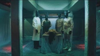 A human body lies on a table between two sets of people wearing dark animal masks and plastic protective coats in a dark, menacing room