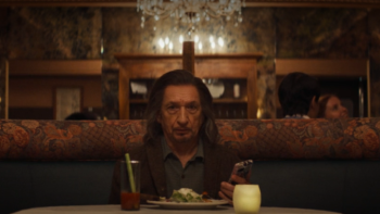 Wonder Man: Trevor sits at a restaurant table with a meal in front of him; he's holding his phone in one hand and appears stunned