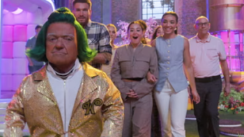 Wonka's The Golden Ticket TV show screenshot 2: A green haired short Oompa-loompa in beige overalls leads contestants down colorful corridor.