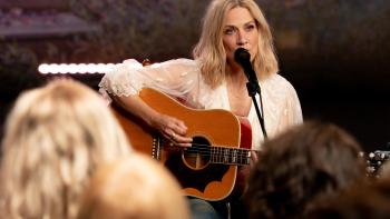 Words + Music TV show screenshot: Sheryl Crow holds a guitar and sits while singing in front of an intimate crowd.