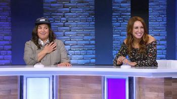 Comedians Sabreena Jalees and Rachel Feinstein sit on a game-show set with a brick backdrop. There's a long paneled desk in front of them, each of them has a buzzer directly in front of them, they are both smiling..