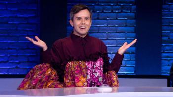 Comedian Cole Escola sits behine a paneled desk on a game show set with a blue brick backdrop. There is a game show buzzer on the desk in front of them. They are smiling with their arms are up in a "Am I right?" type pose, they wear a voluminous dress in shades of maroon, orange, and hot pink.