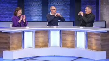 Comedian Matt Walsh sits at a long paneled desk on a game show set, flanked by The New Yorker's Naomi Fry and fashion designer Isaac Mizrahi. Walsh and Mizrahi are making heart shapes with their hands, Fri looks on with a laugh while clapping her hands. 