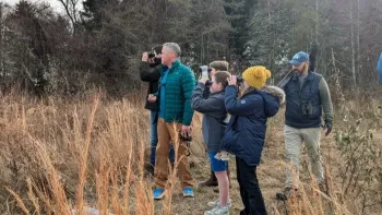 Jeff Corwin and a group of kids and adults all stand bundled up in warm clothes while standing outside looking in the same direction with binoculars