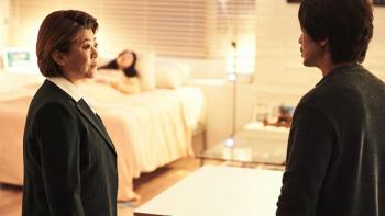 A middle-aged woman in a suit with chin-length hair stands in a bedroom speaking to a man whose face is not shown. In the background, a woman with long, dark hair lays in a bed under a white comforter. An IV pole is visible next to the bed.