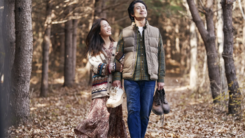A man and woman walking through a forest in autumnal clothing, holding their shoes. The woman holds the man's arm, and they both have beatific expressions.
