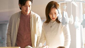 A man and woman stand over a crib with a mobile hanging over it. They look down at a baby, his expression somewhat blank and hers a bit unnerved.