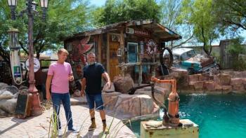 Zillow Gone Wild TV screenshot: Host McBrayer and a homeowner stand near a pool of water, surrounded by auto-themed items.